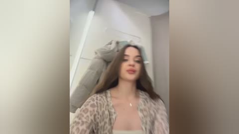 Video of a young woman with long brown hair, wearing a leopard-print cardigan, standing in a dimly lit hallway with beige walls and a door.