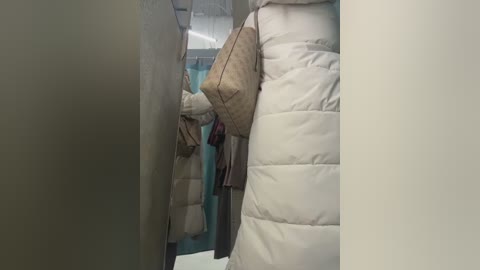 Video of a person in a beige quilted jacket, brown tote bag, and dark pants, leaning against a wall in a narrow, dimly lit hallway with teal-painted walls and a door visible in the background.