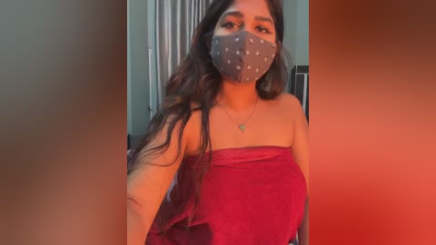 Video of a woman with long, wavy dark hair, wearing a gray mask with white polka dots, a red strapless dress, and a necklace, standing indoors with a red and orange gradient background.