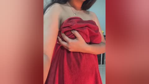 A video of a woman with medium brown skin and long black hair, wrapped in a red towel, standing indoors.