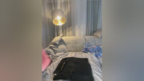 Video of a messy, dimly lit bedroom with a gray couch, scattered blankets, a black bag, and a yellow and gray pendant light hanging from the ceiling.