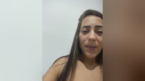 A video of a young woman with long, dark hair, light brown skin, and a neutral expression, partially hidden by a beige wall, wearing a dark top.