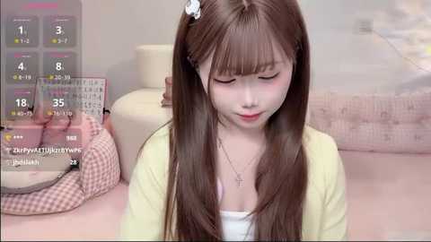 Media: A video of an East Asian girl with long brown hair, wearing a yellow cardigan, sitting on a bed with a pink checkered blanket. The background features a calendar and a beige wall.