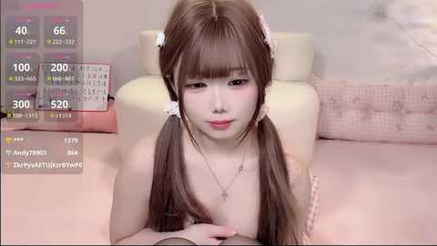 Video of an East Asian woman with long brown hair in pigtails, wearing a pastel pink bra and a necklace, lying on a bed with a light pink sheet.