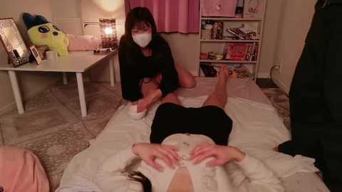 Video of two Asian women in a cozy, dimly lit room. One is massaging another on a bed, wearing a face mask and black outfit. Toys, a pink lamp, and plushies adorn the background.