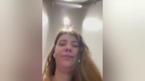 A video of a young woman with long, light brown hair, wearing headphones, looking down with a slight smile. The background features a dimly lit, narrow hallway with white walls and a ceiling light.