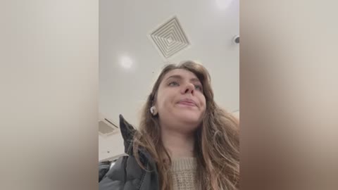 Video of a young woman with long, wavy brown hair, wearing a dark jacket, standing in a narrow, brightly lit hallway with a circular vent on the ceiling.