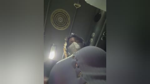 Video of a person in a black jacket and a white face mask, seen from behind, standing in a dimly lit, industrial-looking room with a circular air vent and a large black cylindrical object.