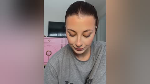 Video of a young woman with light skin and dark hair tied back, wearing a gray shirt, looking down thoughtfully. Background features a pink dresser and a flat-screen TV.