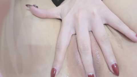 Video of a close-up view of a person's hand with red-painted nails, gently touching their shaved vulva. The skin is light-toned and the background is out of focus, emphasizing the intimate and explicit nature of the image.