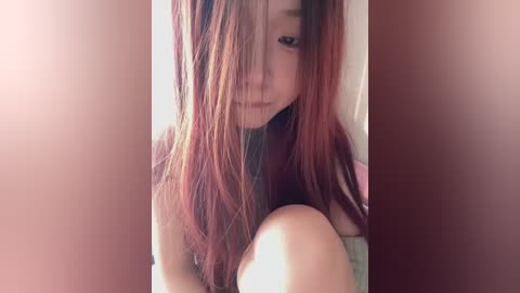 Video of an Asian woman with long, straight, auburn hair, partially covering her face, sitting on a bed with a soft, warm lighting, creating a cozy and intimate atmosphere.