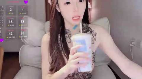 Video of a slender Asian woman with long brown hair, wearing a black lace bra, drinking a blue milkshake, sitting on a light-colored couch, with a digital overlay showing 18, 48, and 82 viewers.