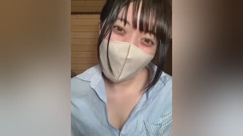 Video of an Asian woman with straight black hair and bangs, wearing a light blue striped shirt, beige face mask, and brown eyes. Background shows a wooden wall.