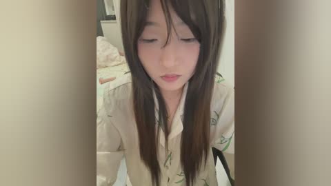 A video of an Asian woman with long, straight black hair, wearing a white shirt with green floral patterns, sitting in a dimly lit room. She has a serene expression, and the background shows a bed with white sheets.