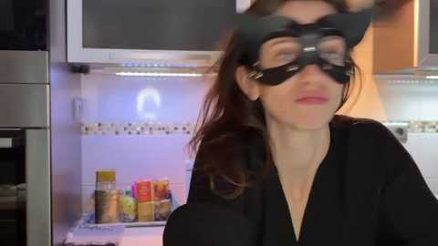 Video of a woman in a black cat mask, with long brown hair, wearing a black robe, standing in a kitchen with open refrigerator displaying spices.