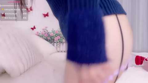 A video featuring a woman with light skin, wearing a blue sweater and black thong, bending over a white couch adorned with red butterflies and flowers, with text overlays indicating a live stream.