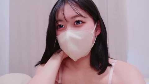 Video of a young Asian woman with straight black hair, light skin, and a pale face mask, wearing a light pink tank top, with blurred background.