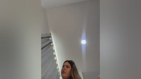 A video of a young woman with long, light brown hair, standing in a modern, minimalist room with white walls and a recessed ceiling light.