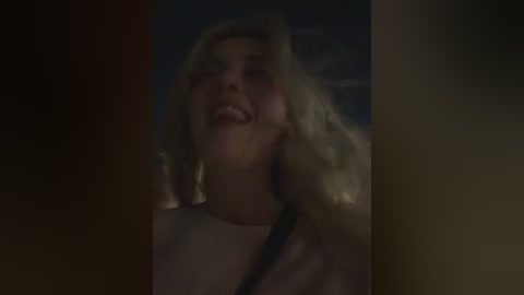 A dimly lit video of a blonde woman with shoulder-length wavy hair, smiling widely, partially obscured by a dark background.