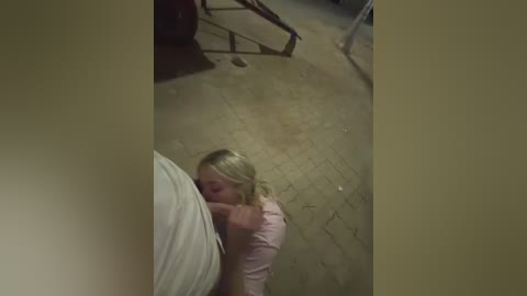 A video captures a young blonde girl, wearing a pink top, kneeling to suck a man's erect penis, which is partially visible. The background shows a dimly lit, cluttered room with a wooden floor and a metal object.