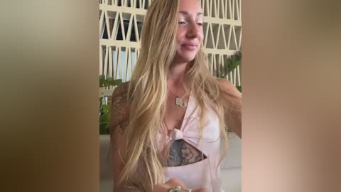 Video of a smiling, long-haired, light-skinned woman with tattoos, wearing a pink halter top, standing indoors with a modern, white geometric wall and green plants in the background.