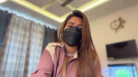 Video of a woman with long, straight brown hair, wearing a black face mask, pink jacket, and large hoop earrings, in a brightly lit room with modern decor and a TV on the wall.