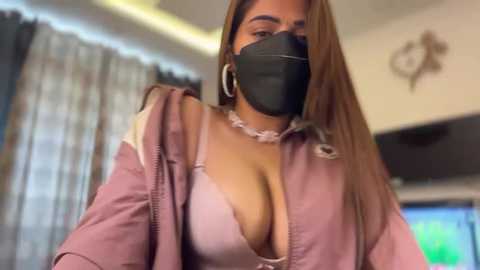 A video of a light-skinned woman with long, straight brown hair, wearing a black mask, pink jacket, and a revealing white bra. She stands in a dimly-lit room with curtains and a TV in the background.