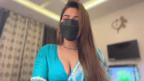 Video of a woman with long brown hair, wearing a black face mask, turquoise top, and a silver choker, in a modern living room with patterned curtains and a TV.