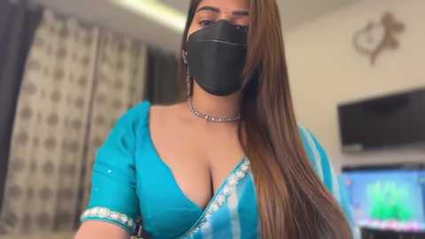 Video of a South Asian woman with long brown hair, wearing a turquoise saree with a deep V-neck, black face mask, and a silver choker. She stands indoors, with curtains, a TV, and a wall-mounted heart decoration in the background.