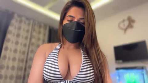Video of a young woman with long brown hair, wearing a black face mask, black and white striped bikini top revealing ample cleavage, in a modern living room with a TV and patterned curtains.
