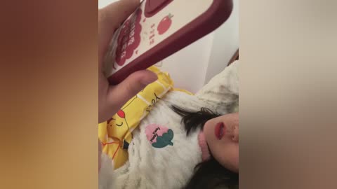 Video of a woman with dark hair, wearing a yellow shirt with cartoon characters, lying on a bed, taking a selfie with a red phone, partially visible.