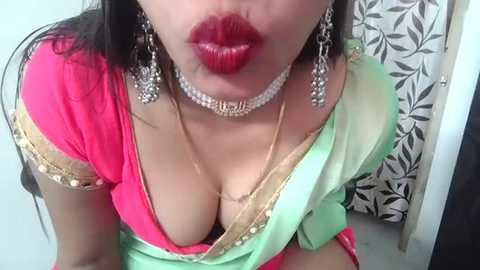 Video of an Indian woman with medium skin tone, wearing a bright pink and green sari with gold embroidery, deep red lipstick, and large silver earrings. She's indoors, with a floral-patterned curtain in the background.