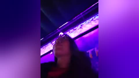 Media: Video of a person with long, dark hair and a black jacket, standing under a purple and blue neon-lit bar ceiling, giving an abstract, surreal atmosphere.