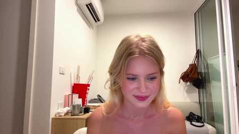 A video of a blonde woman with fair skin and light makeup, smiling, in a minimalistic bathroom with white walls, a red trash can, and a brown handbag hanging.
