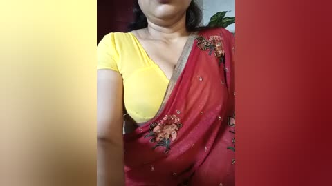 Video of a South Asian woman with medium skin tone, wearing a yellow short-sleeved blouse and a red saree with floral embroidery, against a red and yellow gradient background.