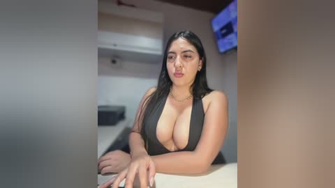 Video of a curvy, Latina woman with long, straight black hair and medium skin tone, wearing a revealing black halter top that accentuates her large breasts. She sits at a white table in a modern, minimalist room with white walls and a TV mounted on the wall.