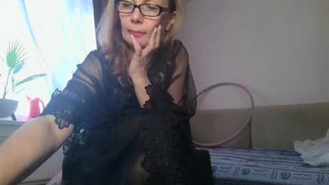 Video of a middle-aged woman with glasses and fair skin, wearing a sheer black lace robe, resting her face on her hand, in a cozy indoor setting with potted plants and a blue-striped cushion.