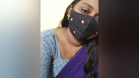 Video of a South Asian woman with medium brown skin, wearing a black face mask with floral designs, a purple saree with gold threads, and a blue lace top, against a beige background.
