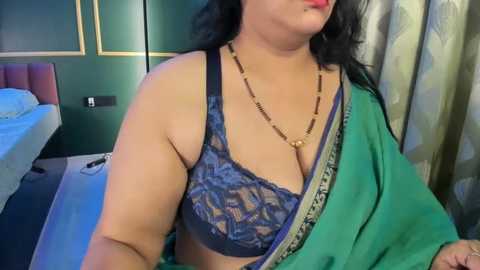 Video of a plus-sized woman with medium skin tone, wearing a green sari and a blue lace bra, standing in a modern bedroom with green walls and a white bed.