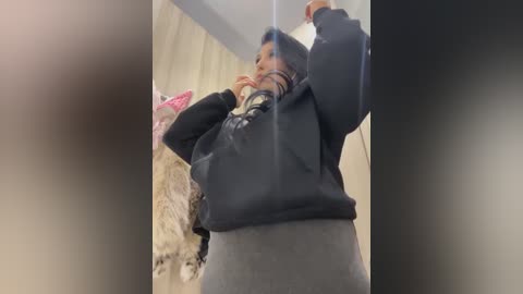 Video of a person with a medium build, wearing a black hoodie and grey pants, holding a phone while adjusting a pink hair accessory, set in a dimly lit room with beige curtains and a fluffy white pillow.