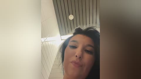 Video of a young woman with long black hair, wearing heavy makeup, puckering her lips in a bathroom with white tiled walls and a wooden ceiling.