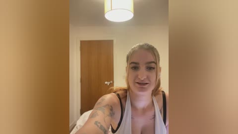 Video of a young, light-skinned woman with blonde hair, smiling, wearing a white tank top, showing tattoos on her arms, standing in a dimly lit hallway with a wooden door and a hanging light fixture in the background.