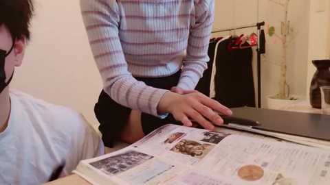 Video of a person in a striped sweater and black skirt reading a magazine at a table in a softly lit, minimalistic room with white walls and hanging clothes.