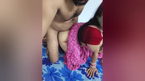 A video shows a nude man with a trimmed beard and a hairy chest, kneeling behind a woman with long black hair and a red dress, who is bent over a blue bedspread adorned with large pink flowers.