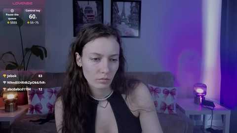 Video of a young woman with long brown hair, wearing a black halter top, sitting on a couch with a floral-patterned blanket. Background includes a lit candle, purple lighting, and framed cityscape pictures.