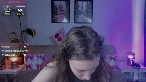 Video of a woman with long brown hair, wearing a black top, sitting on a bed with pink and white patterned pillows. The room has purple lighting and framed pictures of Paris on the wall.
