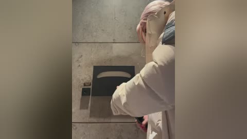 A video of a person in a beige winter jacket, with pink hair, peering into a public restroom urinal with a blue and white design. The background shows tiled walls and a blurry, beige door frame.
