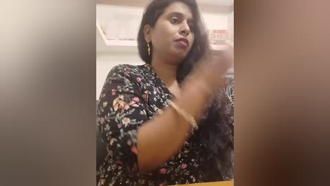Video of a South Asian woman with medium skin tone, long black hair, and a floral-patterned blouse, looking slightly to the side, blurred background.