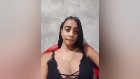 Video of a young woman with medium-brown skin, long black hair, and a black lace bra with crisscross straps, sitting against a textured gray wall. She has a relaxed, neutral expression.