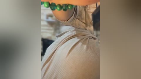 A close-up video of a woman's upper body wearing a grey, ribbed turtleneck sweater, showcasing her ample breasts and a green beaded necklace. The background is blurred and indistinct.